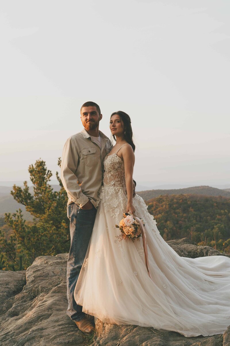 A bride in a flowing wedding gown holds a bouquet, standing on a rocky ledge with scenic mountains in the background.