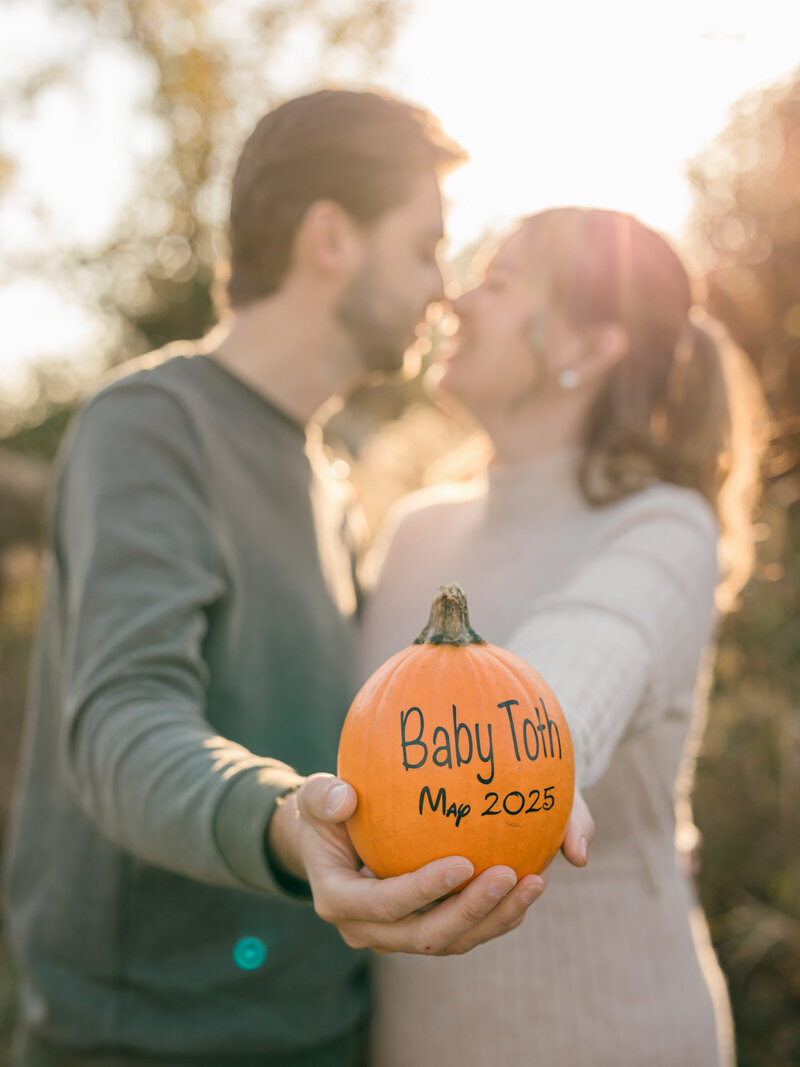 A couple shares a sweet moment in warm golden sunlight while holding a small pumpkin with 'Baby Due' written on it, announcing their pregnancy. The soft autumn glow and natural outdoor setting create a cozy and heartwarming scene. Photographed by KCP Families.