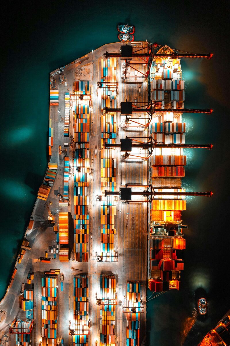 An aerial view of a brightly lit cargo port with numerous colorful shipping containers and cranes, surrounded by dark blue water.