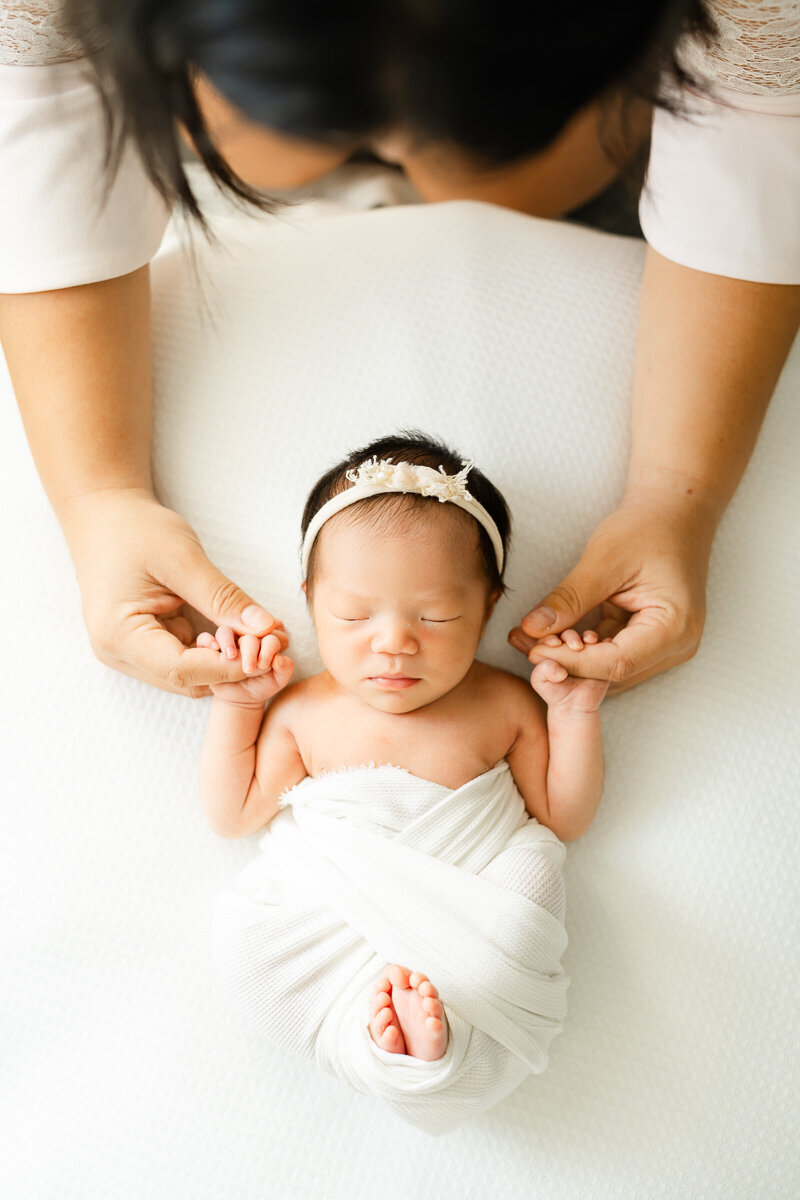 a sleeping newborn girl lays on a white bean bag wrapped in a white swaddle while her mother leans over and holds her hands during their Austin studio newborn session.