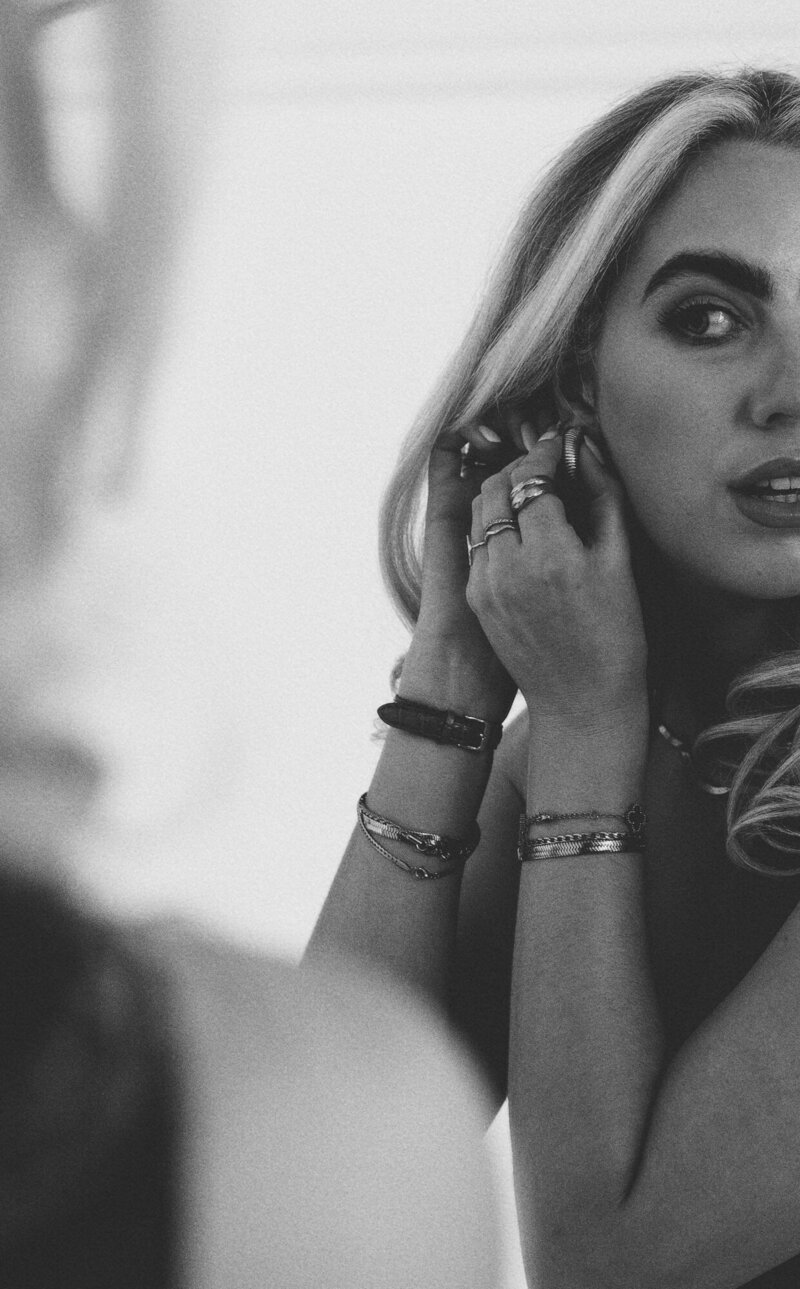 Black and white close-up of Amelia Waters adjusting her earring with layered jewelry and loose curls