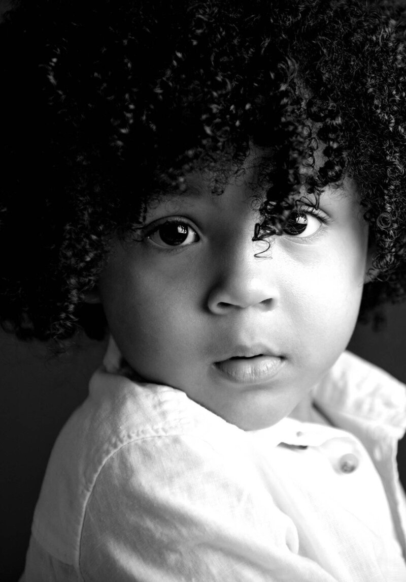 A black and white photo of a young child with curly hair, smiling and looking playfully at the camera.
