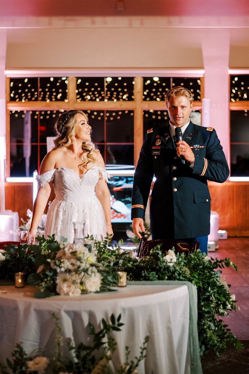 A bride in a white dress and a groom in a military uniform stand smiling at a wedding reception table, with string lights in the background. This scene is part of our full service wedding planning package.