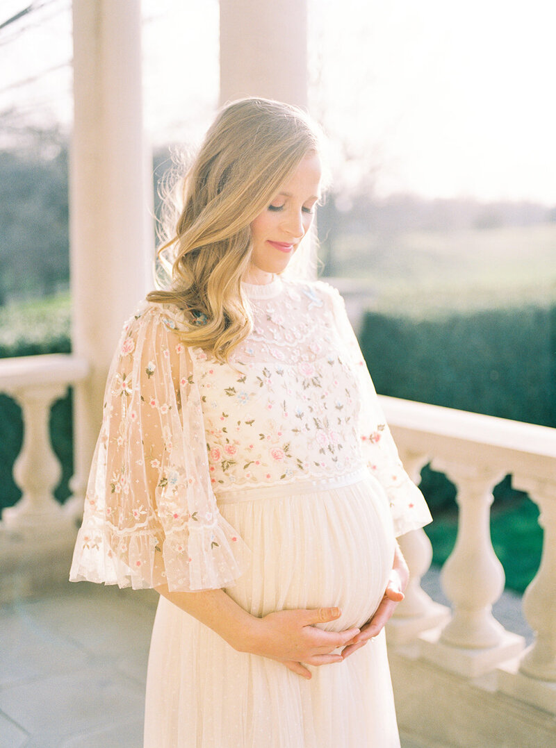 Blonde pregnant mother in ivory Needle & Thread dress stands on balcony with hands underneath her belly.