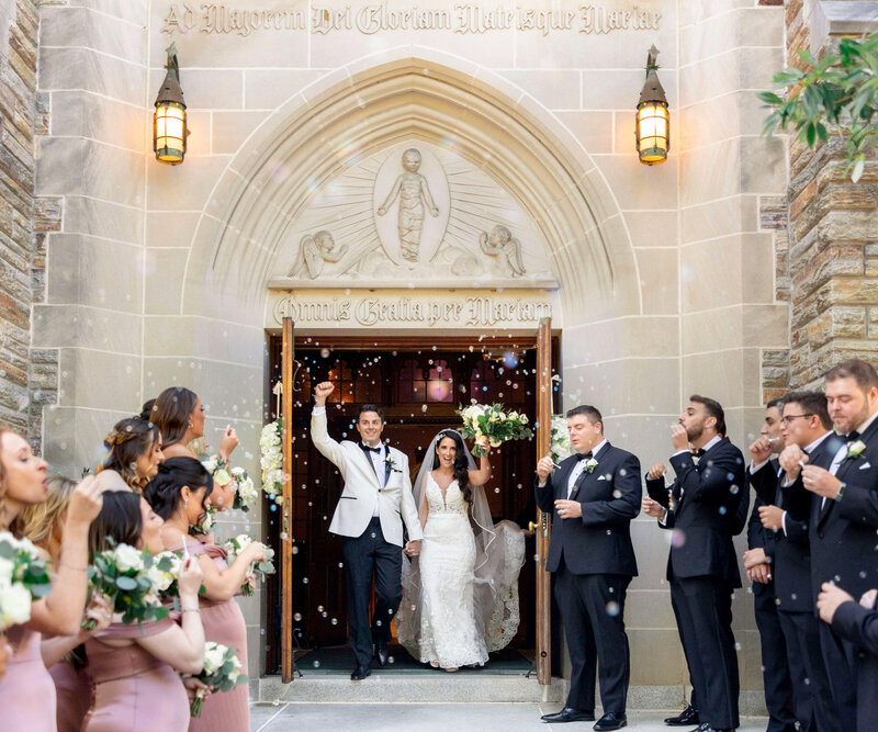 A bride and groom exit a church, smiling and holding hands. The groom holds up his fist in celebration. Guests stand on either side, blowing bubbles and clapping. Everyone is dressed in formal attire.