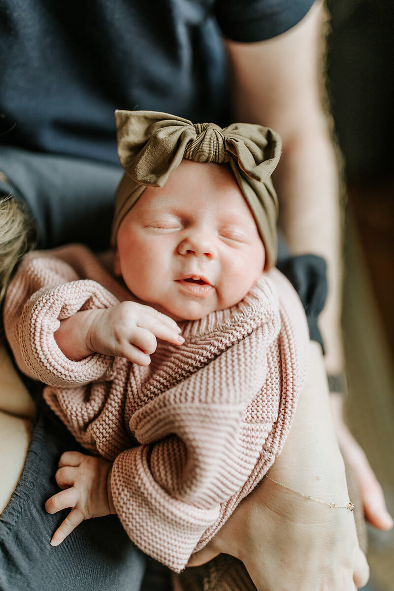 Newborn baby with green bow and cozy mauve sweater held in mom and dad's arms.