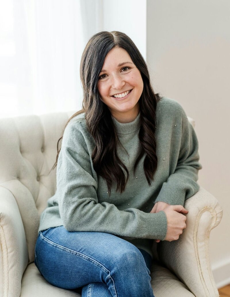 Woman with long dark hair and a warm smile sitting in a cozy chair, wearing a green sweater and blue jeans