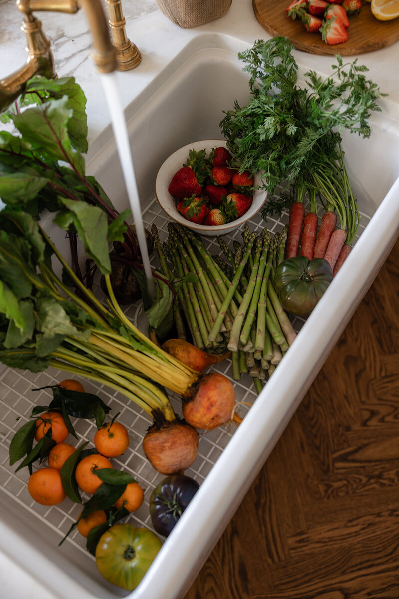 Fresh produce being rinsed in a white farmhouse sink with running water, representing clean, intentional eating
