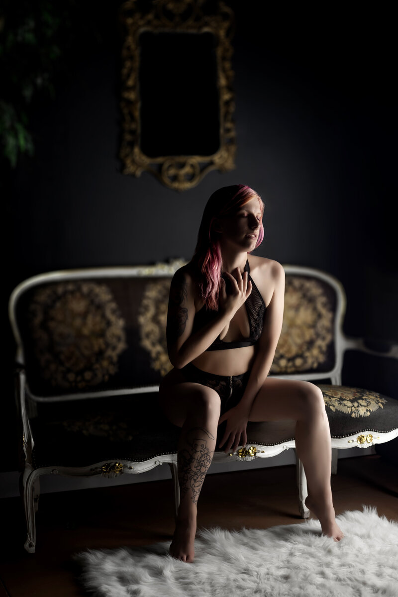 Boudoir photo of woman named Jana in black lace lingerie, sitting on edge of a couch with knees apart, on tiptoes, confidently pulling on her bra strap — photographed by Joclyn in Cedar Rapids, Iowa
