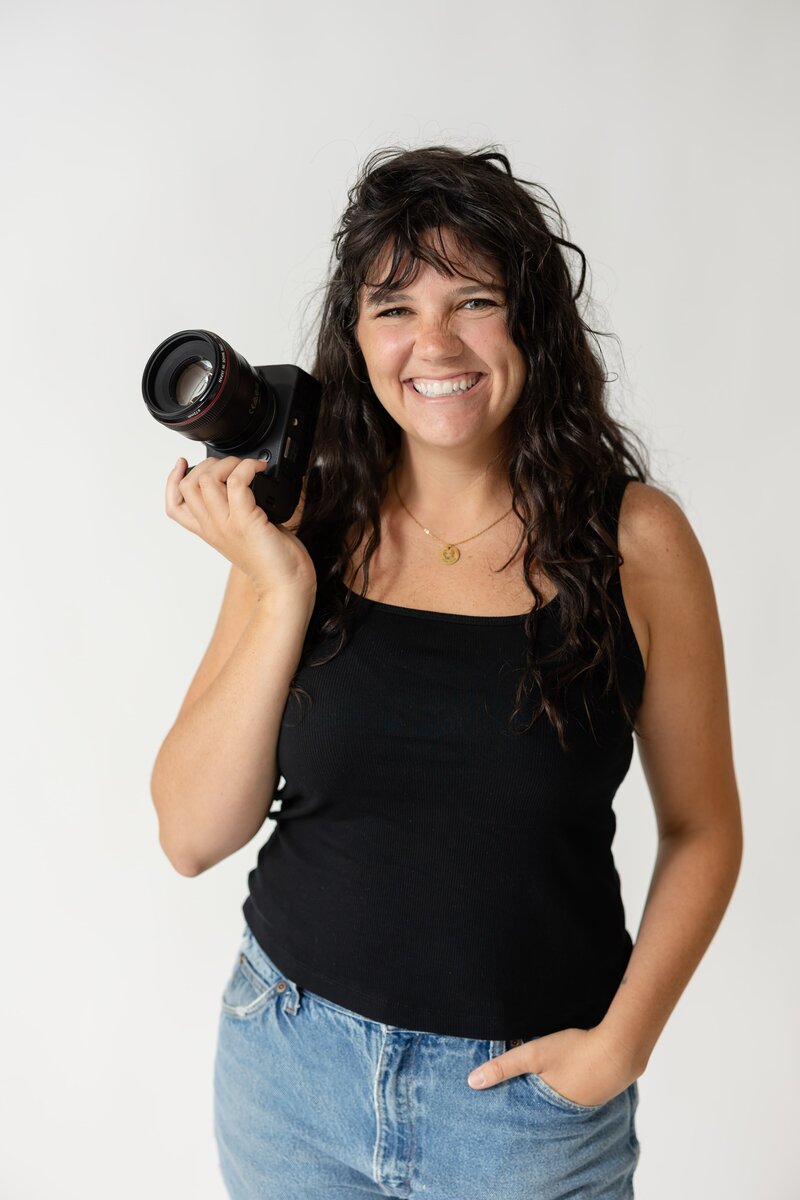 Charleston, SC brand photographer in a studio posing with her camera. She has dark hair and bangs and is wearing a black tank top.