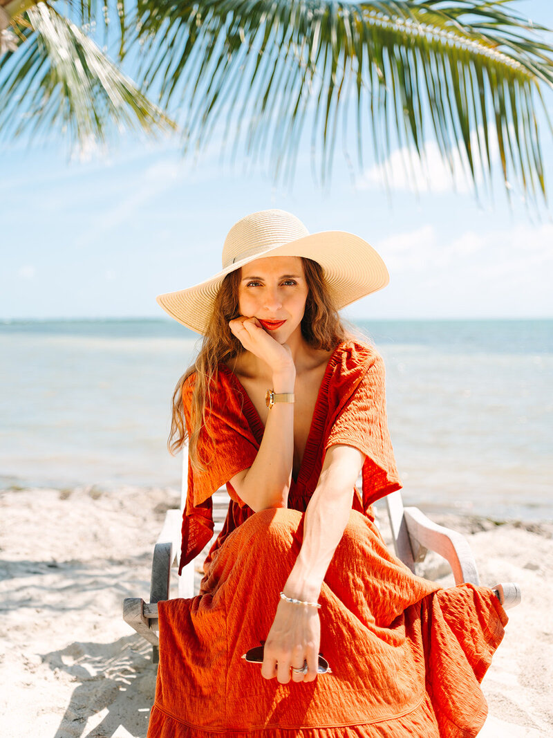 Kilee in an orange dress and straw hat sitting in chair on the beach with ocean behind her