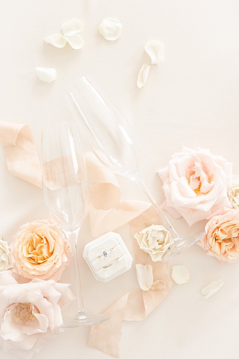 champagne flutes, wedding rings in a ring box, and pastel pink blooms styled with ribbon on a light pink backdrop.