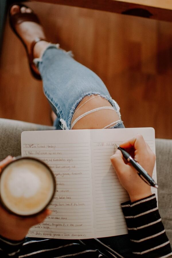 Person wearing ripped jeans and holding a cup of coffee while journaling in a lined notebook, seated in a cozy, casual setting.