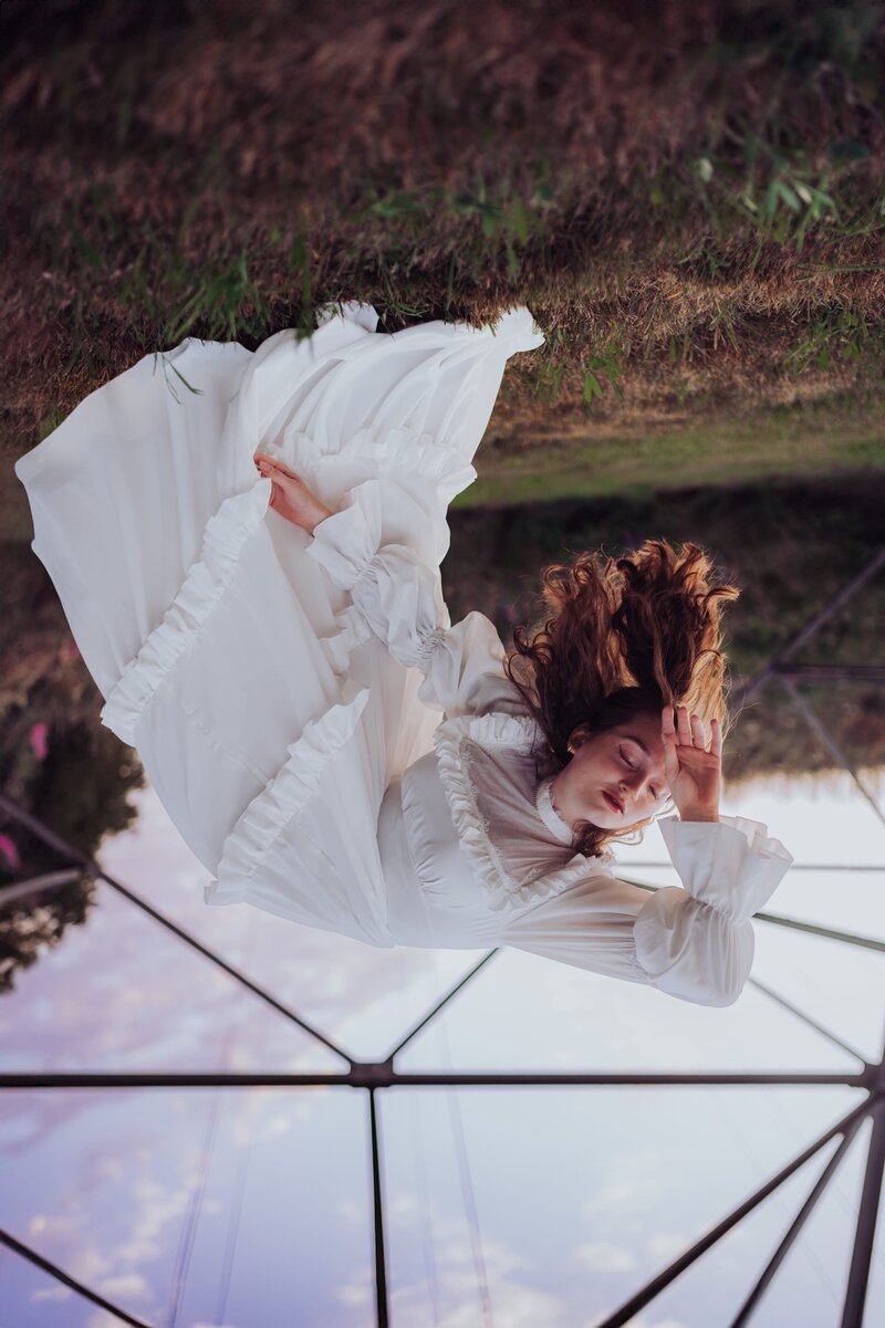 Upside down photo of red-headed woman in white, victorian gown in grassy field with her eyes closed and hand stretched across forehead.