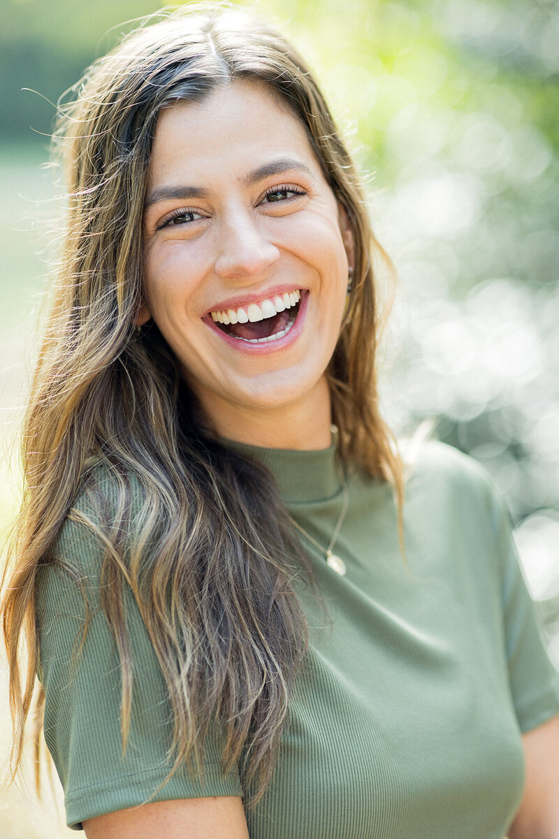 Bright, natural-light portrait of a smiling woman in an olive green top, laughing with a joyful expression. Sunlight filters through the background, creating a soft, bokeh effect. Small business branding photography in Columbia, SC, capturing warm, approachable personal branding for entrepreneurs and professionals.