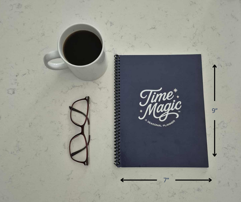 A navy blue Time Magic Planner placed on a light countertop with measurements showing 7 inches wide by 9 inches tall, next to a mug of coffee and a pair of glasses.