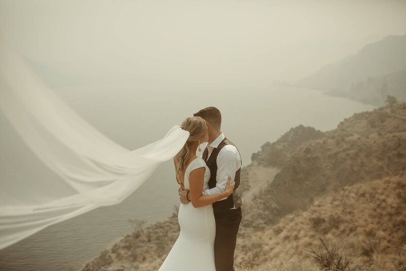 Bride and groom embracing on a coast edge with bridal veil blowing in the wind