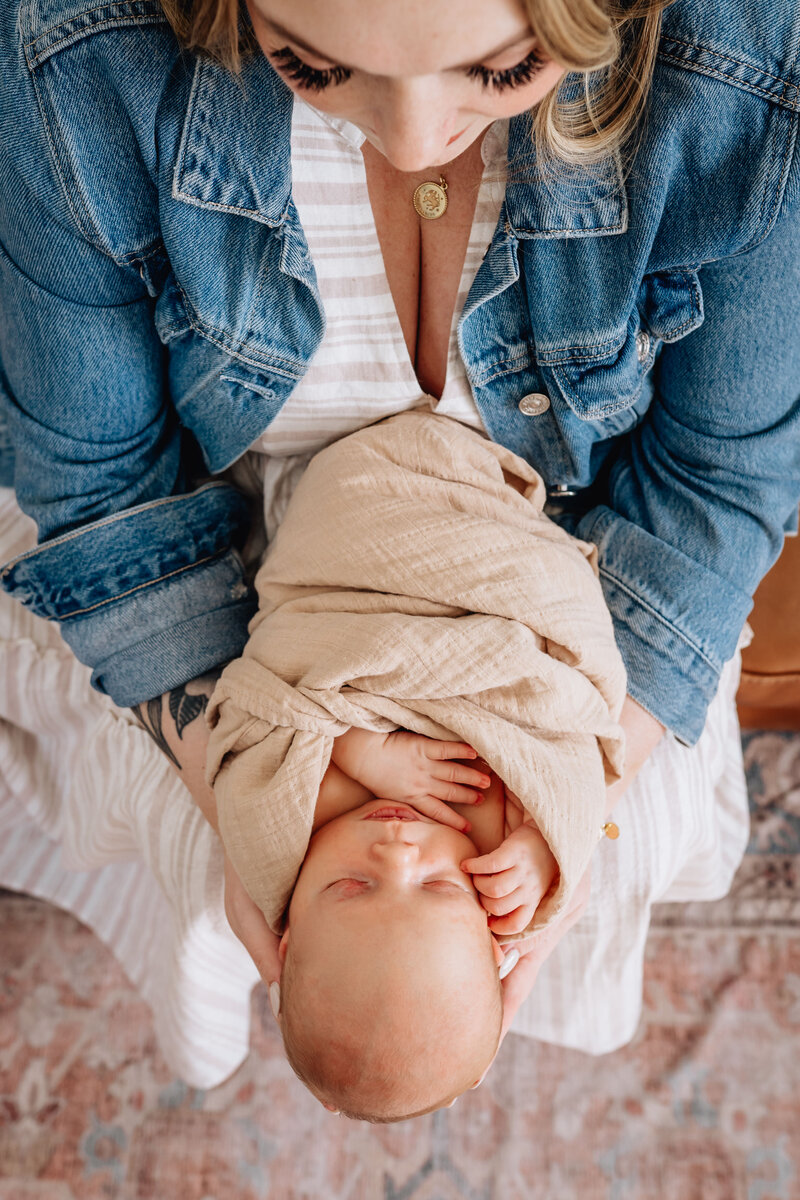 An overhead view of a mother in a denim jacket cradling her swaddled newborn baby. The baby is wrapped in a soft beige blanket, peacefully sleeping as the mother looks down lovingly.