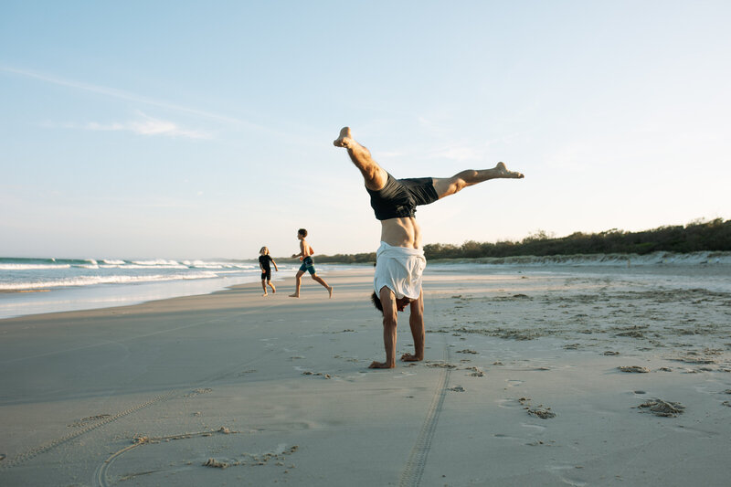  A fun candid moment on a beautiful tweed coast beach, the dad is doing a handstand in the glowing sunset and the boys are chasing each other into the waves