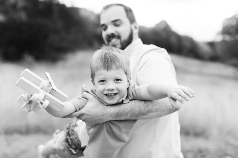 Dad swings his son like an airplane as son holds wooden airplane and pretends to fly