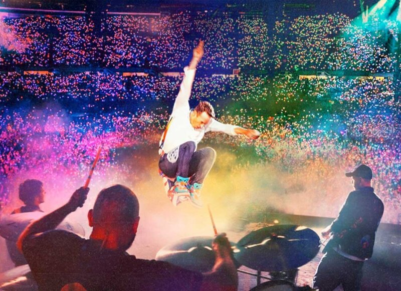 Coldplay lead singer jumps mid-air during stadium concert surrounded by rainbow lights and cheering fans