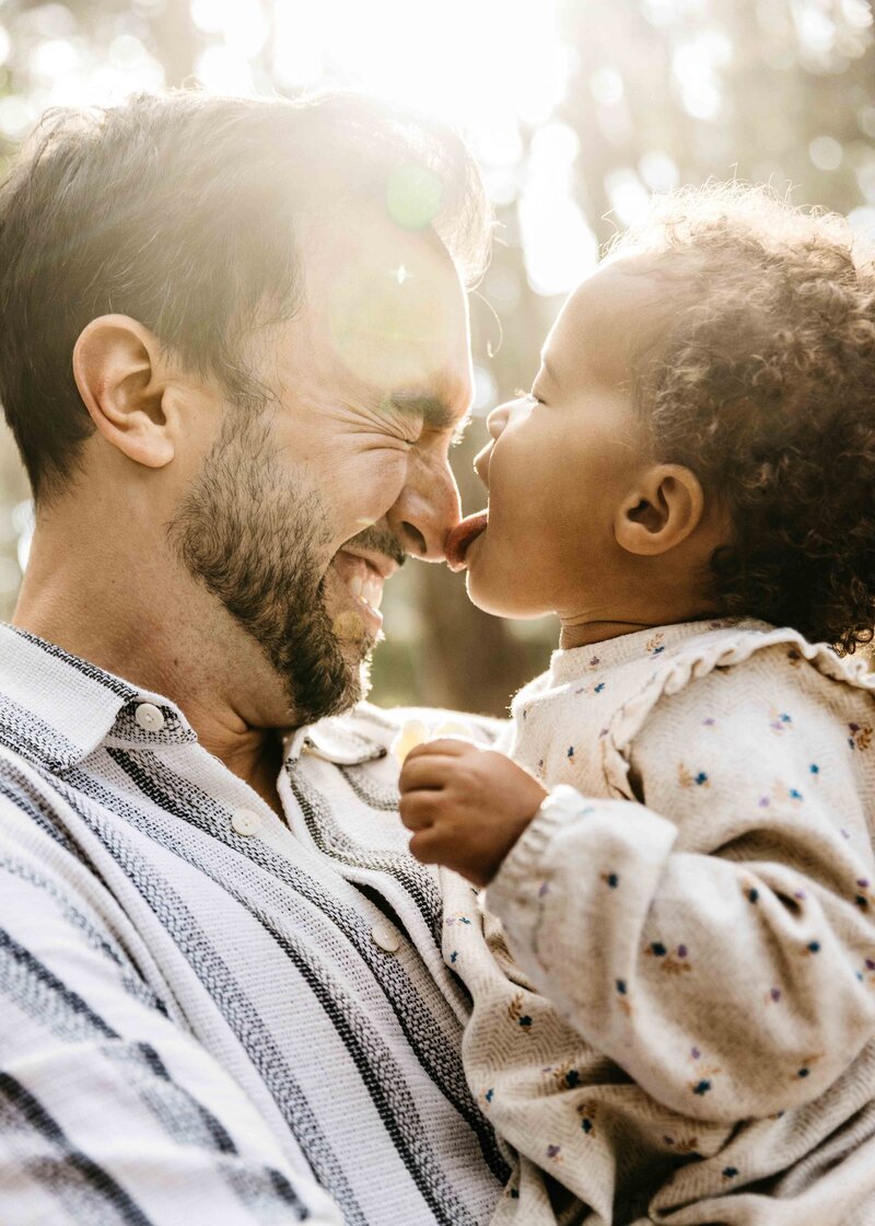 Dad is laughing while carrying his toddler daughter as she licks his nose. 