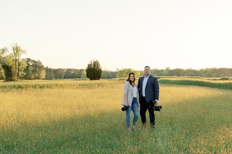 A couple standing in a field with luxury wedding photography equipment, smiling at the camera.