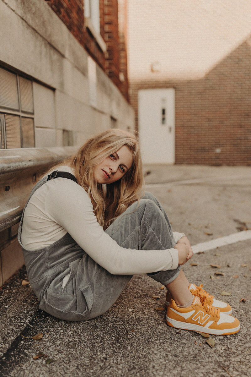 Senior girl in orange nikes and overalls sitting in an alley.