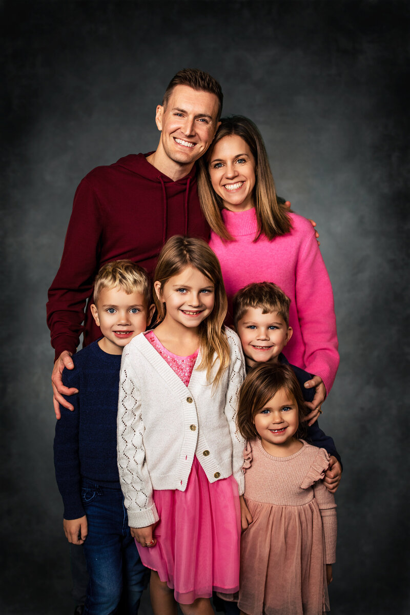 A family portrait of a father and mother and their 4 blonde children pose on a vintage grey studio background.