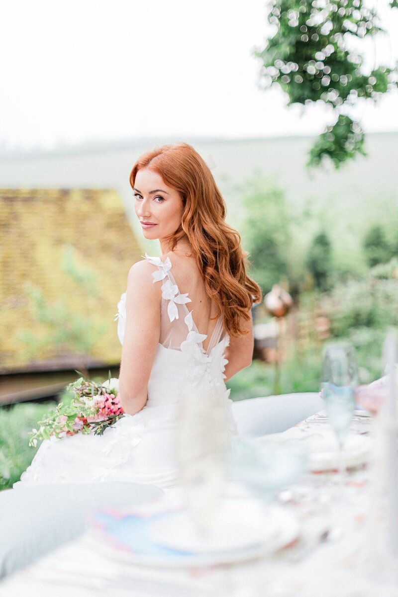 Red-haired bride in a floral appliqué gown turning back with a soft smile during an outdoor editorial-style wedding setup