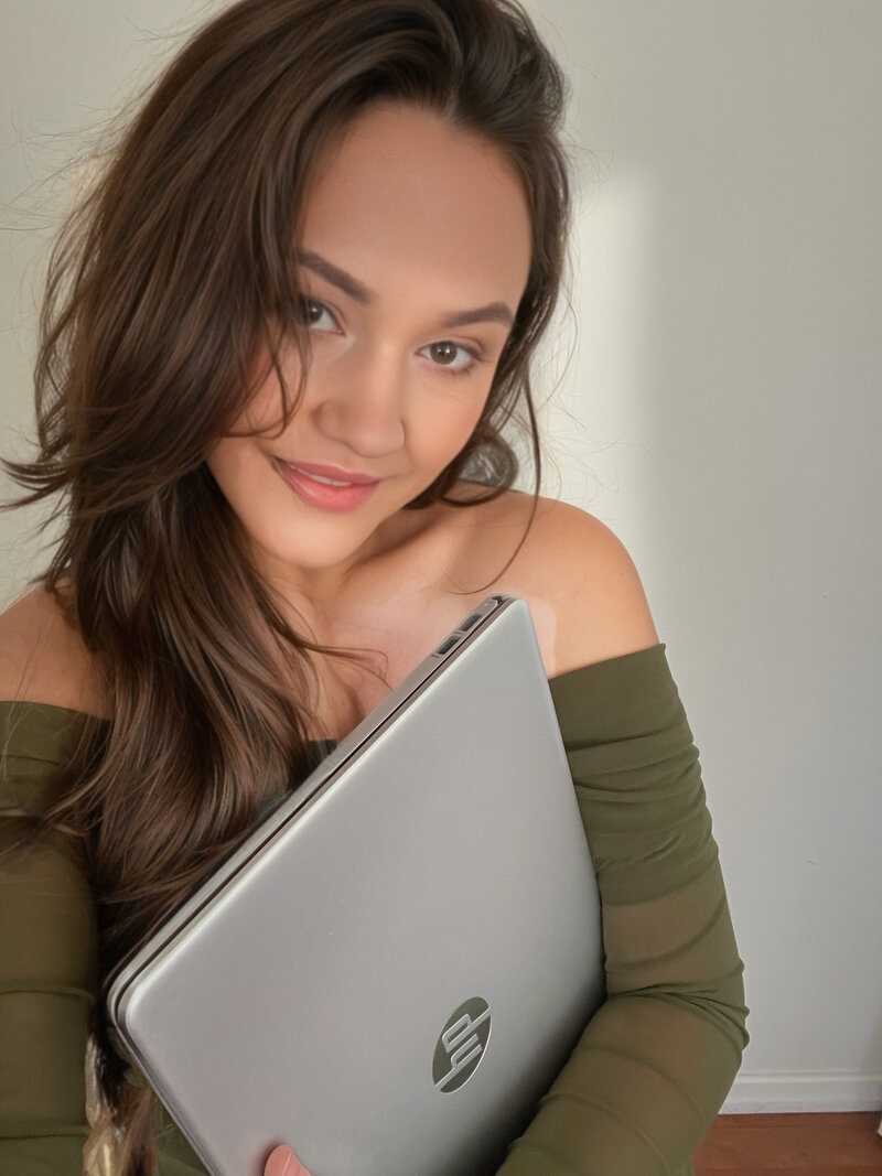 a brunette woman looking at the camera holding a laptop in her arms smiling