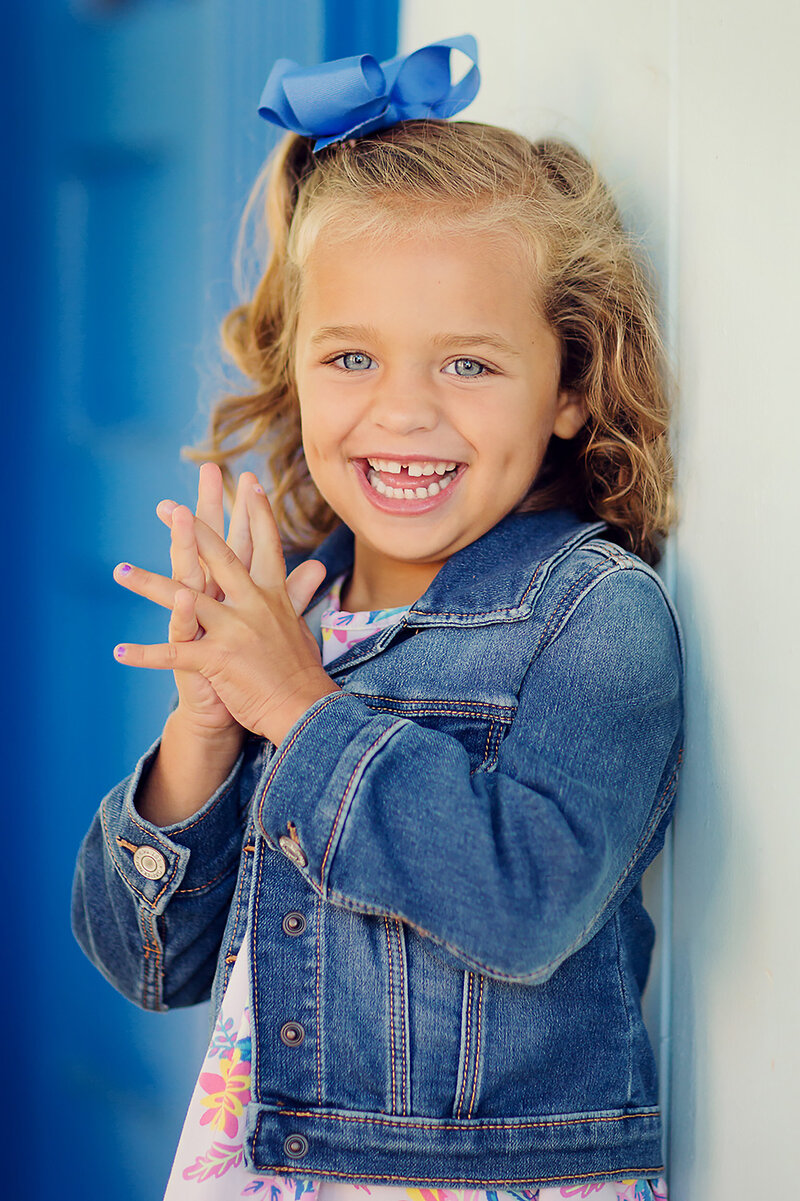 Young girl in a denim jacket and blue bow, smiling very big at the camera and showing off her dimple.