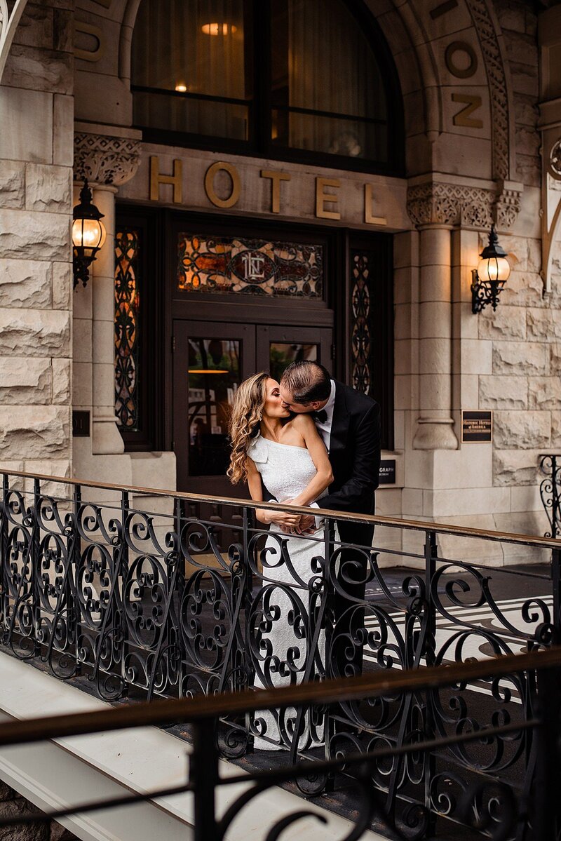 The groom embraces the bride from behind and she turns towards him for a kiss. The bride is wearing  a floor length one shoulder sheath dress with a ruffle on the shoulder. The groom is wearing a black tuxedo with a white shirt and black bow tie. They are standing at a case iron filigree railing in front of a rough hewn stone entry to the Union Station Hotel. The word Hotel is above their heads in gold lettering with two sconces lit up on either side. The door to the historic Nashville hotel featured stained glass transoms and windows set into the dark wood frame.