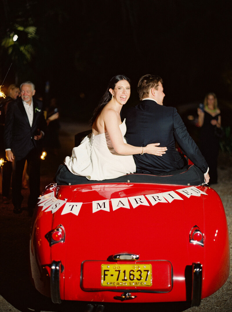 Chic bride and groom drive away from their wedding reception in a red vintage convertible car