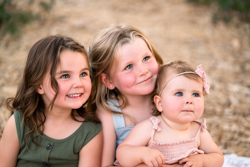 two young sisters holding their baby sister. All have blue or green eyes and are looking off camera