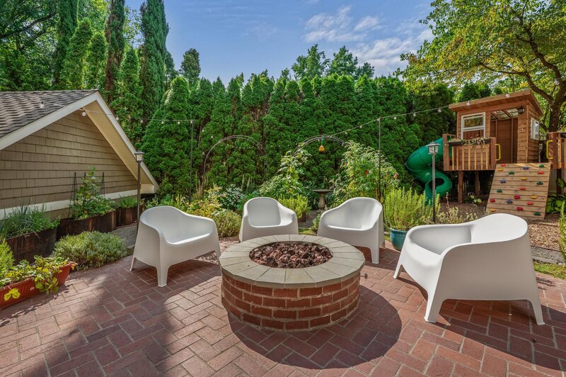 A backyard featuring a brick fire pit with with white chairs surrounding it.