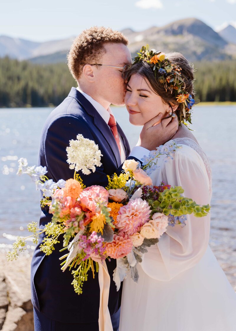 Groom kissing bride’s forehead during a fall elopement at Brainard Lake in Colorado, with colorful bouquet and mountain views in the background