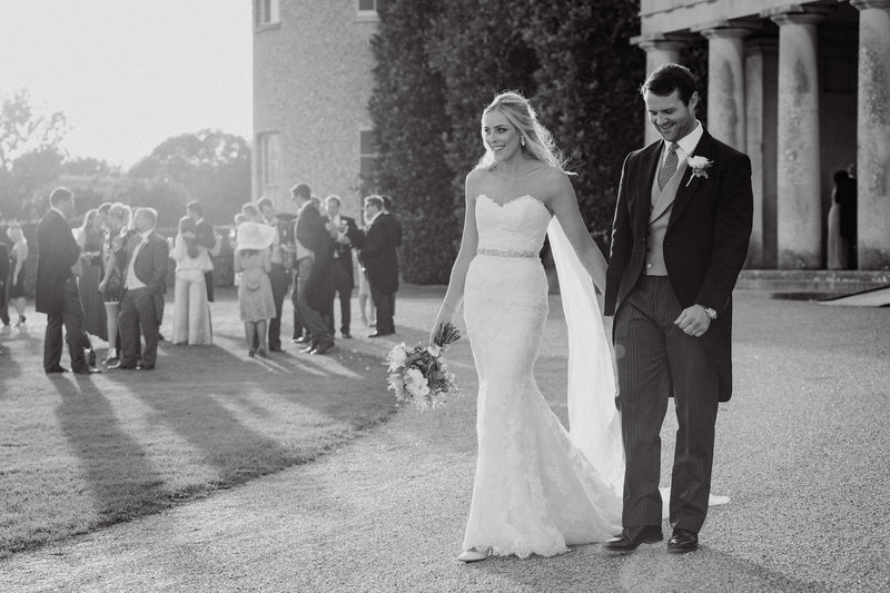 Bride and groom backlight by the sun during golden hour walking hand in hand away from Goodwood House wedding venue in Chichester