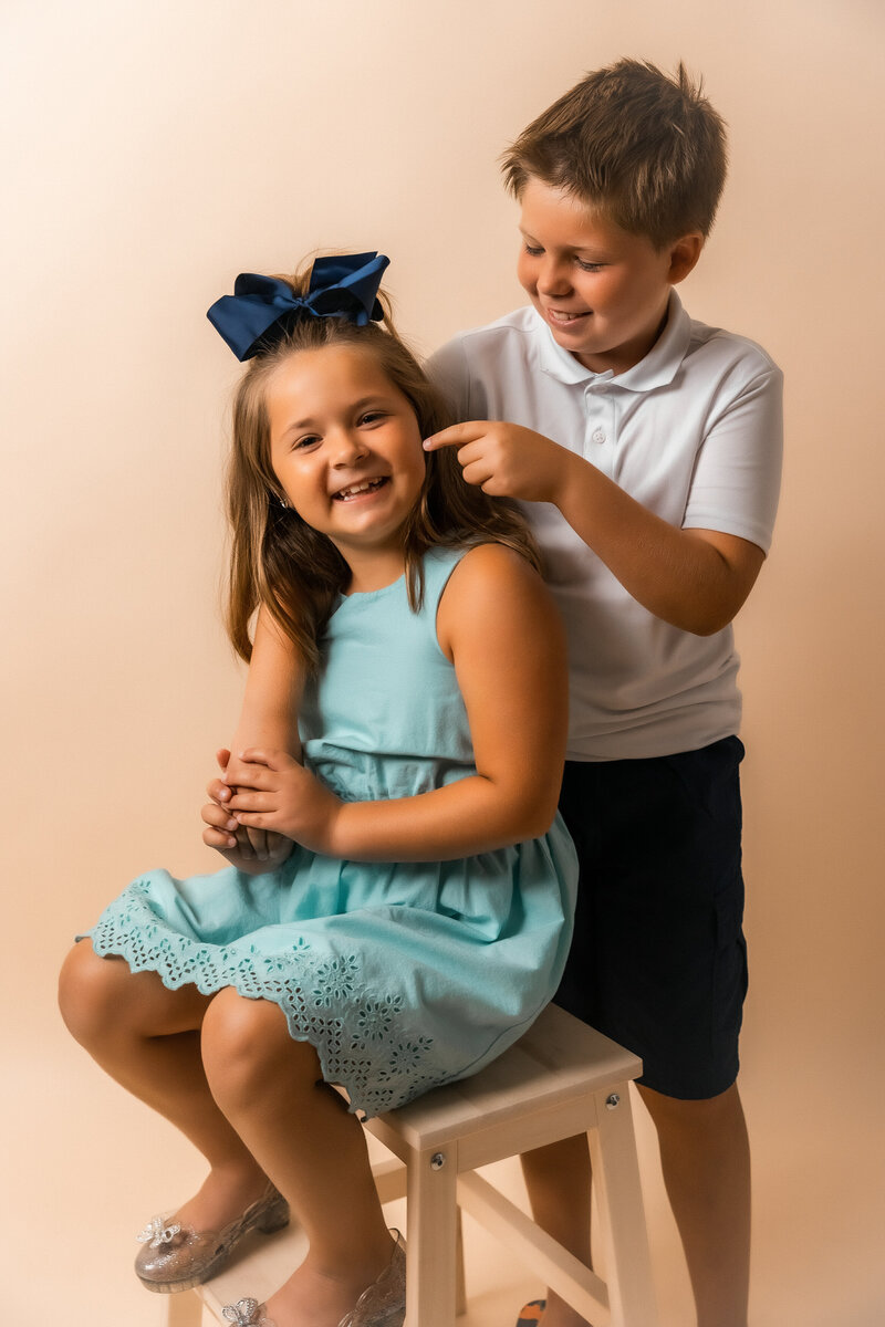 Candid portrait of sister sitting on a stool while brother stands pointing at the sister