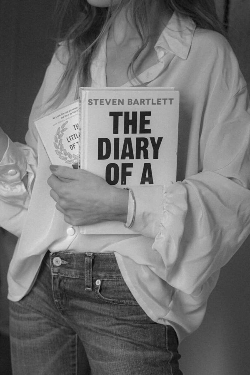 A person in a loose blouse and jeans, perhaps a brand strategist from Sweet Pea Ink Creative, holds two books—one titled "The Diary of a" by Steven Bartlett—in a striking black-and-white photo.