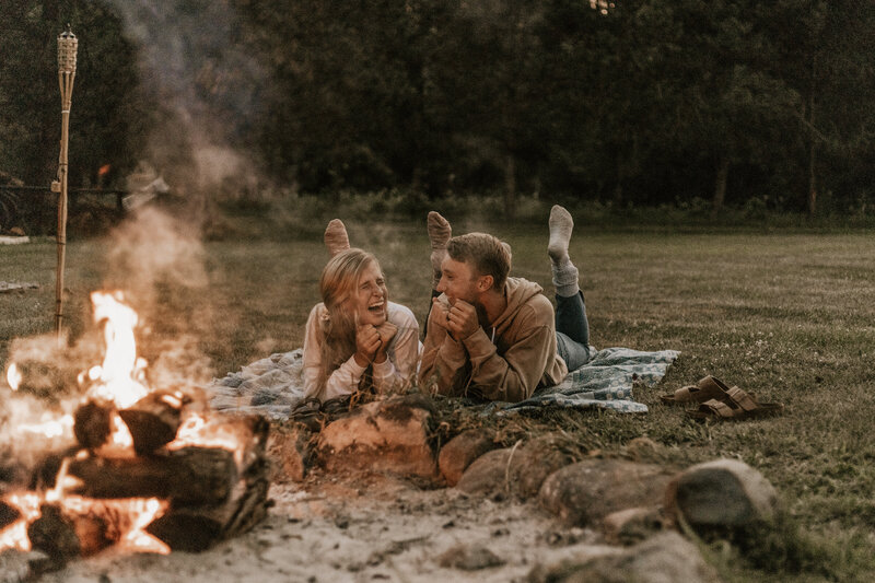 A couple laying on their elbows and laughing in front of a campfire