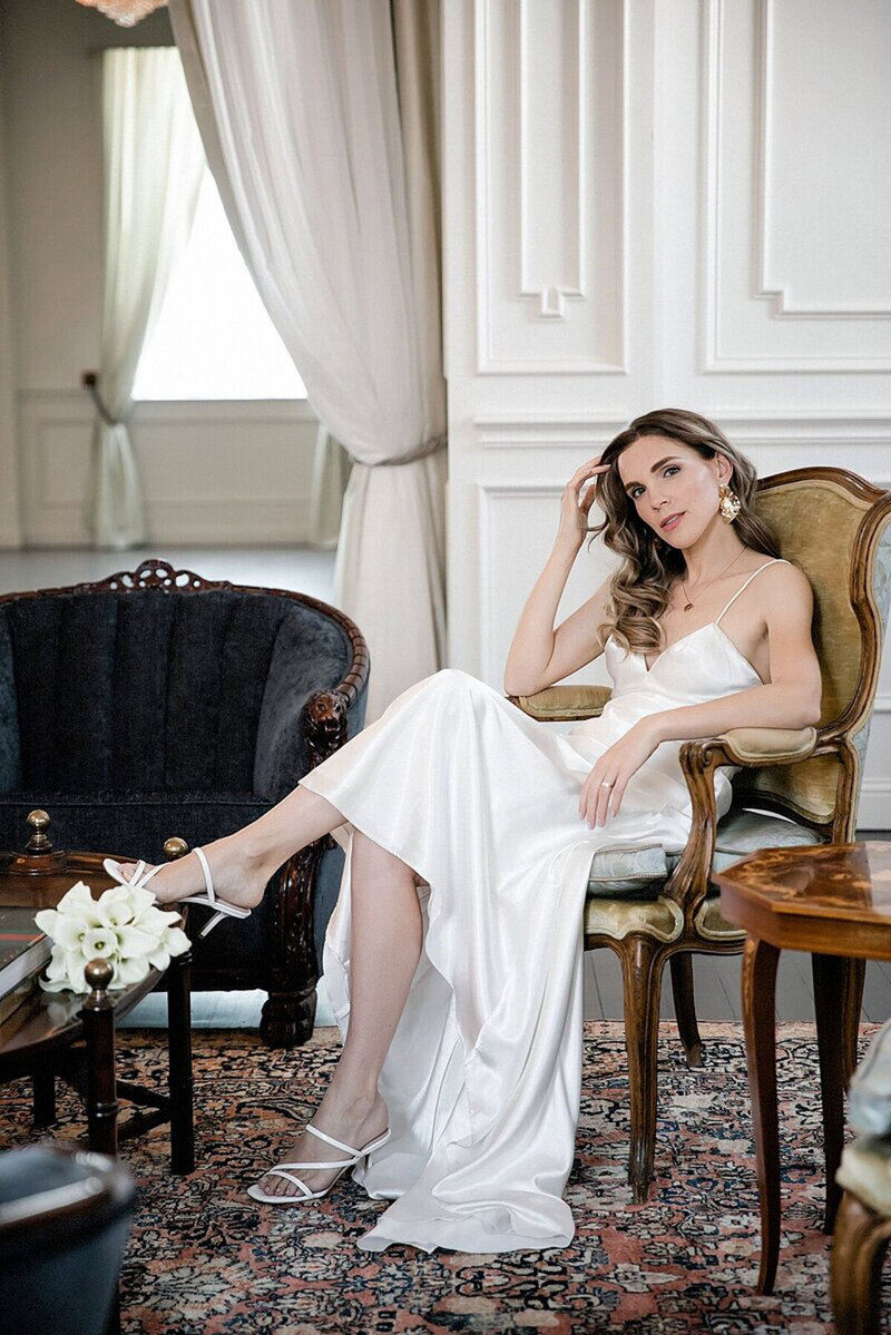 Bride Anna in a silk wedding gown holding a modern white calla lily bouquet, seated in a velvet chair on the second floor of The Mason wedding venue in Dallas.
