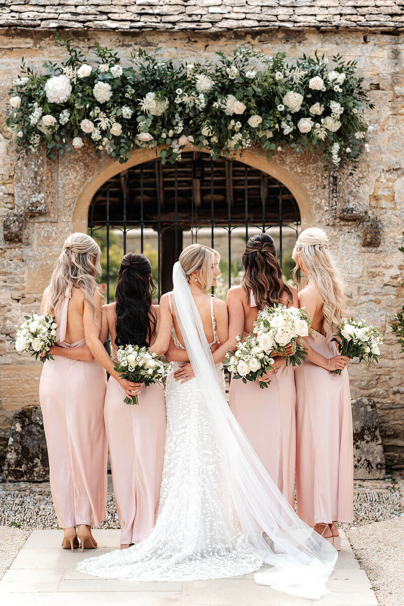 Bride and bridesmaids in soft pink dresses posing outside Caswell House with bouquets