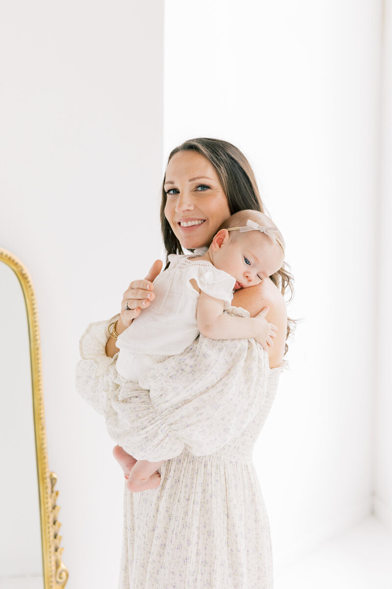 An infant girl falling asleep on mom's shoulder while she smiles and stands in a studio