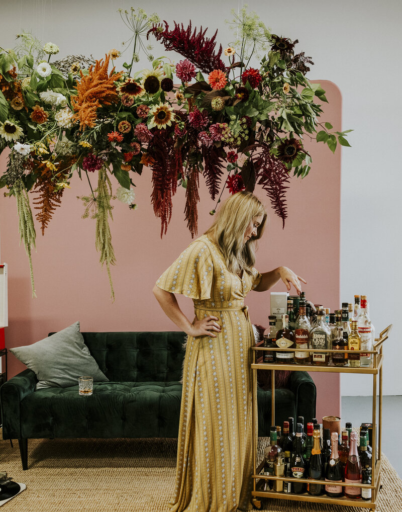 Brittany Frid wears a long 70's patterned yellow maxi dress stands in front of a full gold bar cart in front of a large suspended floral installation with garden flowers, a green velvet couch and a pink backdrop.