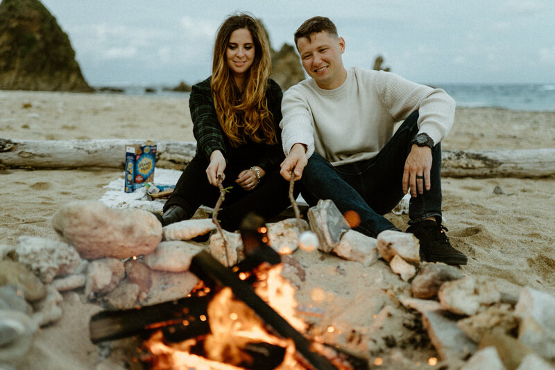 B & T smiling looking at their s'mores cooking over the campfire on the beach