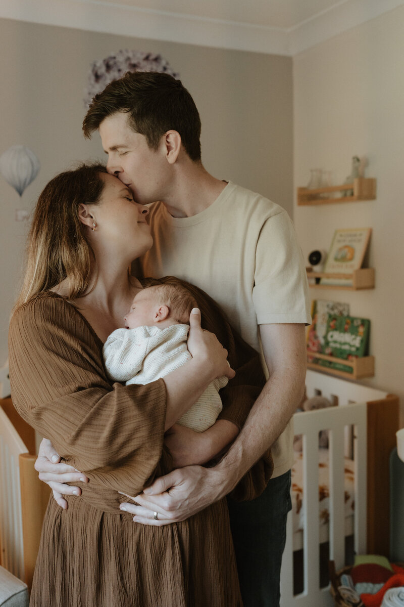 A Brisbane couple gently holds a sleeping baby wrapped in a knitted blanket.