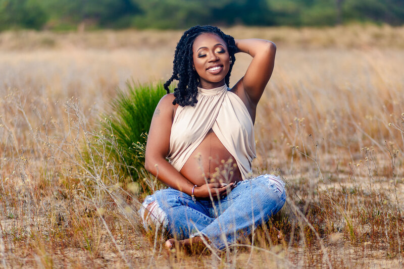A stunning Black mother smiles in a field, proudly showcasing her exposed pregnancy belly and beautiful locs. A radiant celebration of motherhood and natural beauty.