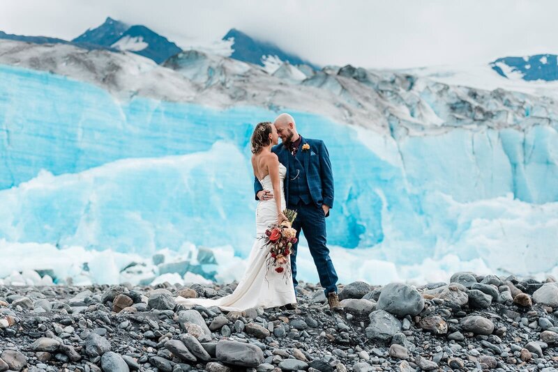 A bride and groom are seated on a dogsled being pulled by a team of sleddogs captured by their Luxury Destination Wedding Photographer during their Alaska elopement. in front of them spreads a snowy glacier field, bright blue skies with snow capped mountains in front and around them peeking out of the glacier.