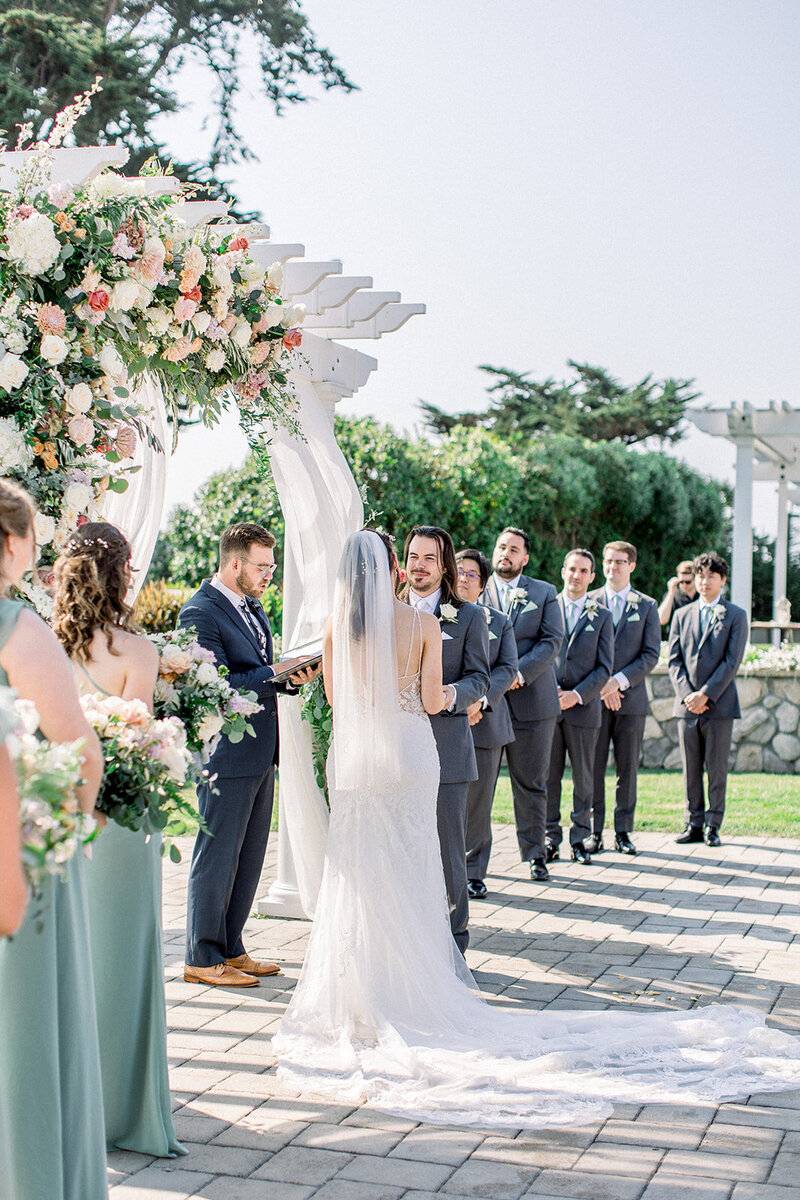 This stunning California wedding ceremony captures the heartfelt moment the couple says their vows beneath a beautifully draped pergola adorned with lush florals. Set against a picturesque backdrop of serene landscapes, the couple exchanges promises surrounded by their wedding party and loved ones. The bright sunlight enhances the elegance of the scene, bringing a soft glow to the bride’s flowing veil and lace gown. Perfectly capturing the timeless beauty of a California wedding celebration.